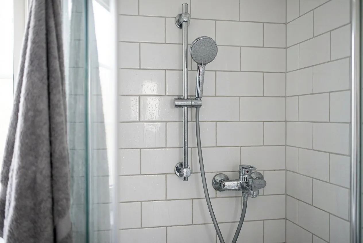 Symbolbild: verchromte Brausestange mit Handbrause und Schlauch an glänzenden weißen Wandfliesen mit Wassertropfen, links unscharf ein Handtuch